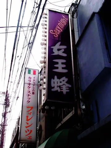 A tall purple Japanese sign rises beside dense overhead cables above a narrow urban lane.