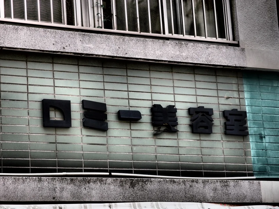 Raised dark salon lettering stretches across pale green rectangular tiles beneath a barred upper window.
