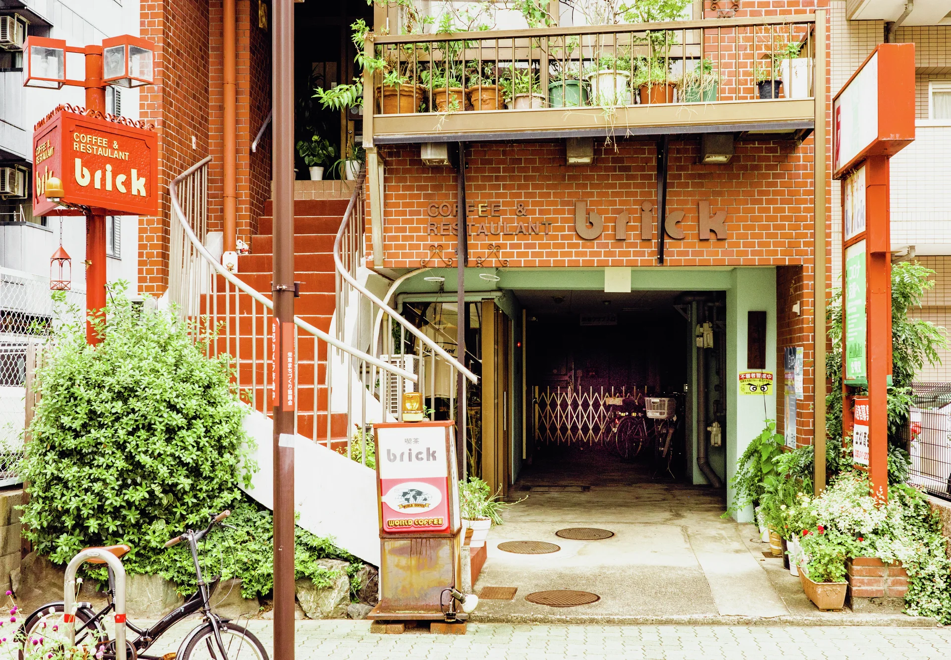A brick building has an exterior staircase, plant-filled balcony, and recessed entrance beside a bicycle.