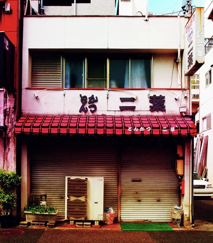 A red tiled awning shelters closed restaurant shutters beside an air conditioner and small pots.