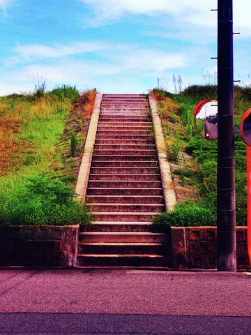 A straight brick stairway rises through grassy banks beneath blue sky beside a dark pole.