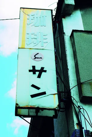 A tall pale turquoise projecting sign with Japanese lettering hangs beside a weathered building wall.