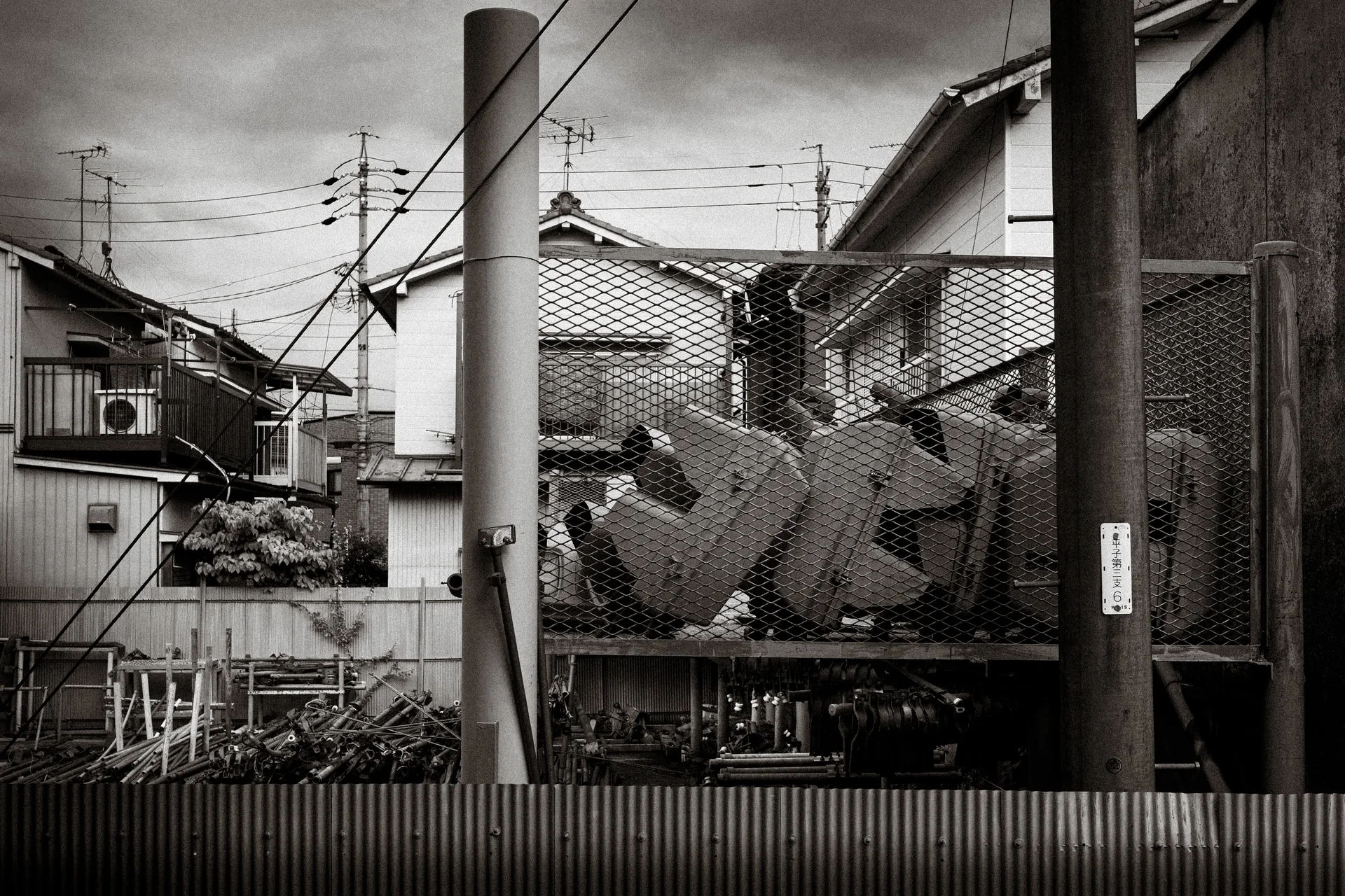 Oversized sign characters sit behind wire mesh between utility poles and tiled rooftops in black and white.