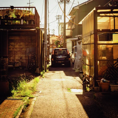 Sunlight floods a narrow alley between weathered buildings leading toward a parked dark van.