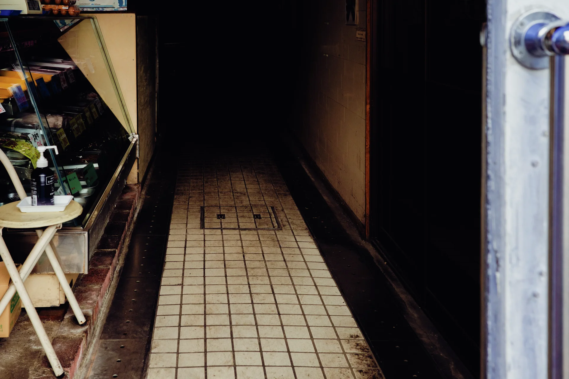 A narrow tiled corridor recedes into darkness beside a cluttered shop display and open metal door.
