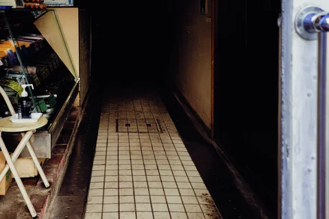 A narrow tiled corridor recedes into darkness beside a cluttered shop display and open metal door.