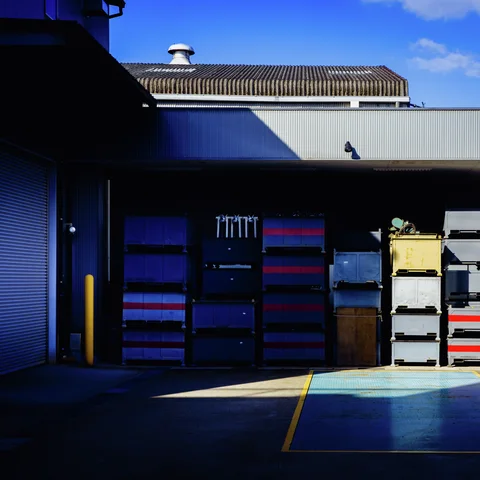 Stacks of blue, gray and red containers line a shadowy loading area beneath a corrugated canopy.