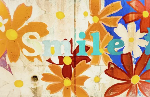 Large orange and white painted flowers surround multicolored Smile lettering on a weathered panel.