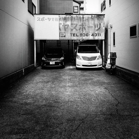 Two cars sit beneath a faded parking sign at the end of a narrow driveway in black and white.