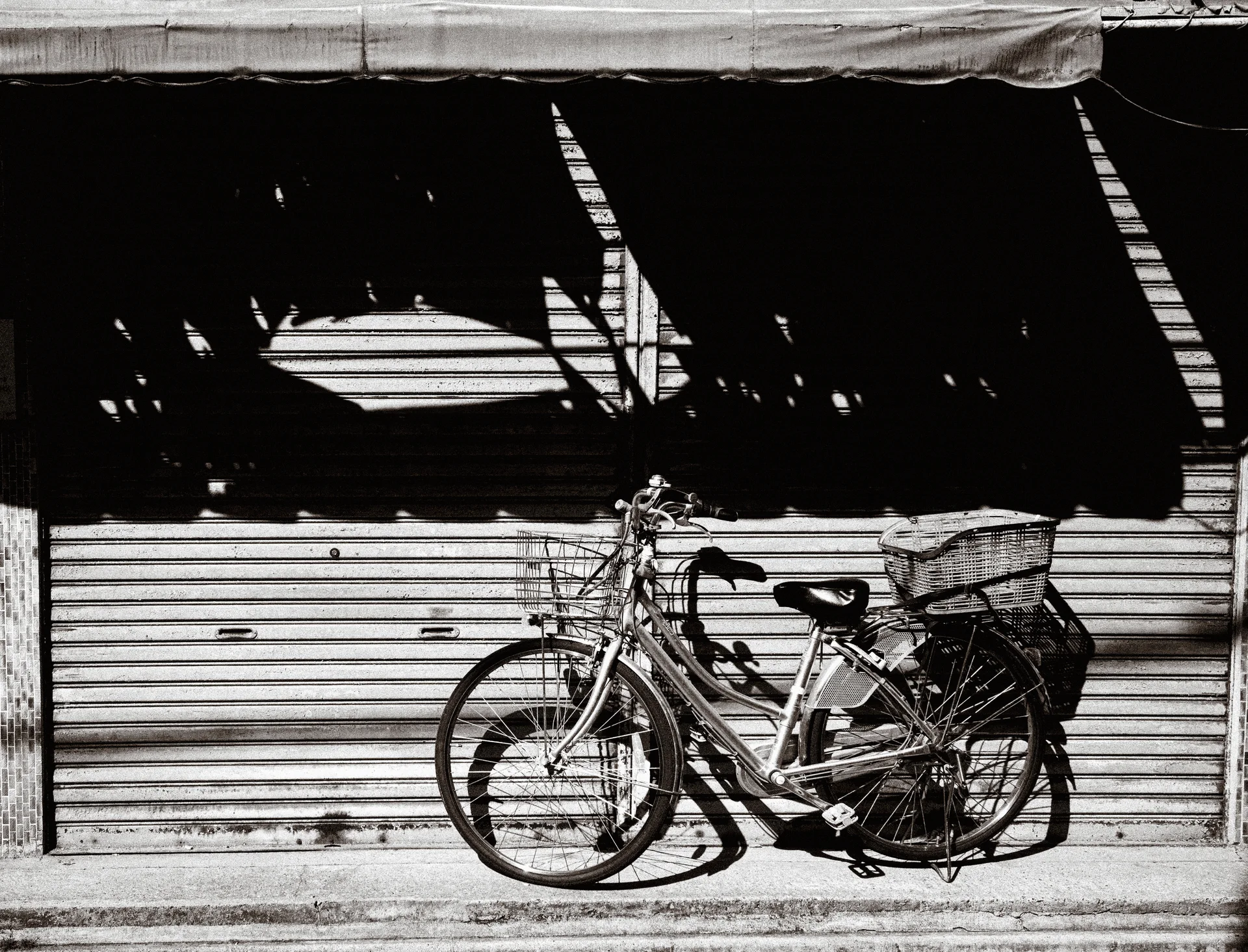 Two bicycles lean against a shutter beneath sharp striped awning shadows in black and white.