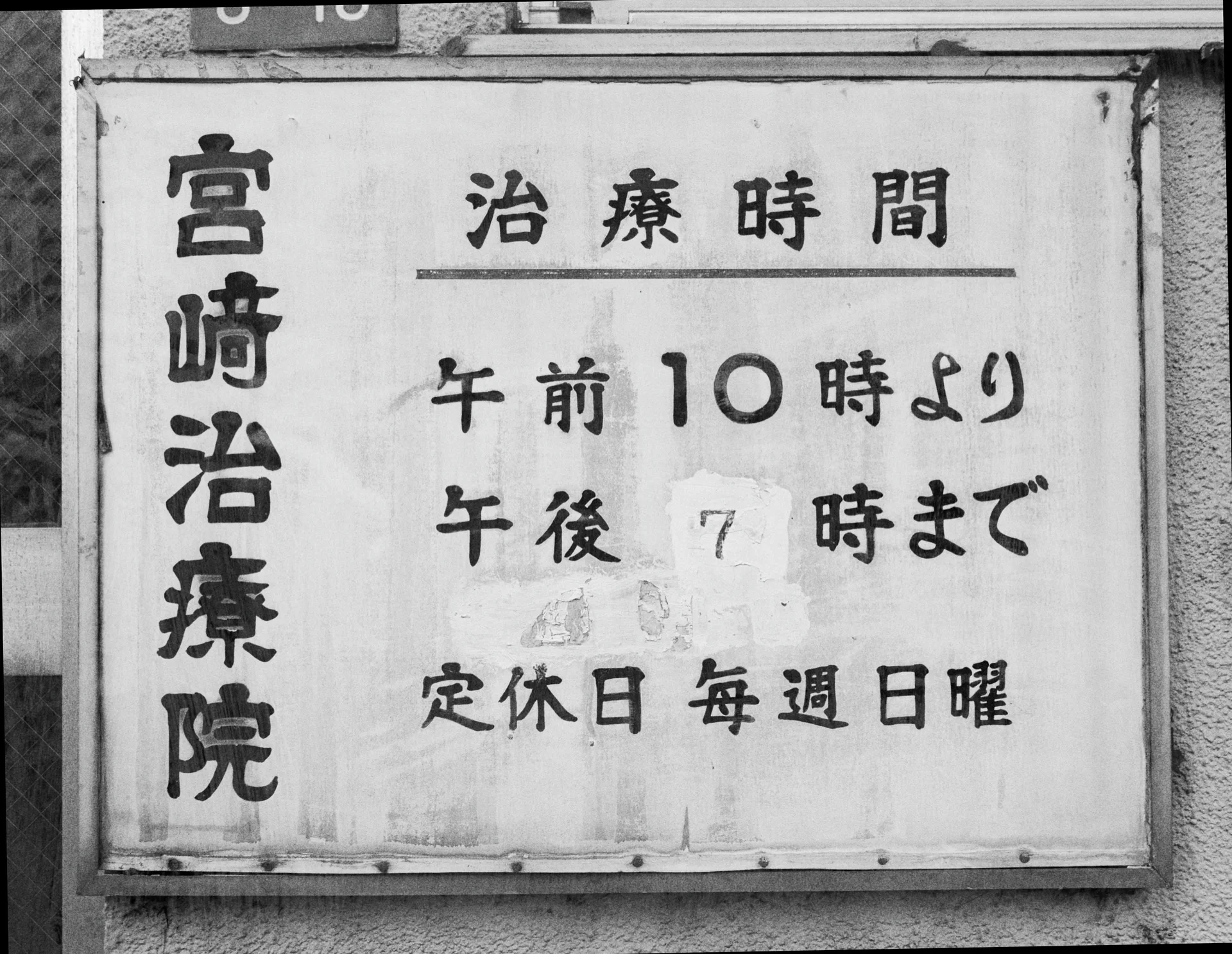 Handwritten Japanese characters cover a weathered rectangular business-hours sign mounted on a wall in black and white.