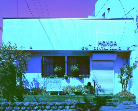 A blue-toned low clinic building has barred windows, a closed shutter, and a planted stone-edged frontage.