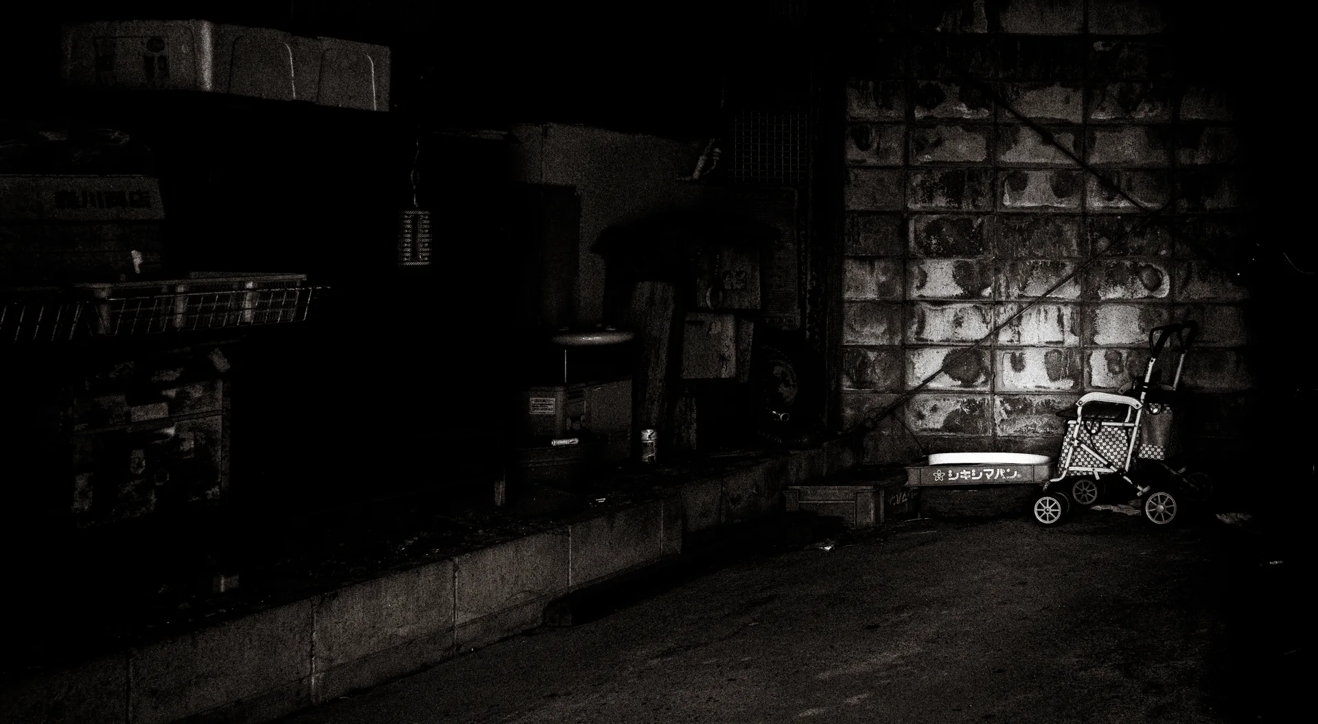 A small wheeled shopping trolley stands beside a sunlit block wall in a deeply shadowed passage.