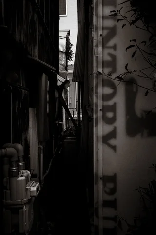 Pipes and faded vertical lettering flank a dark narrow gap between buildings in black and white.