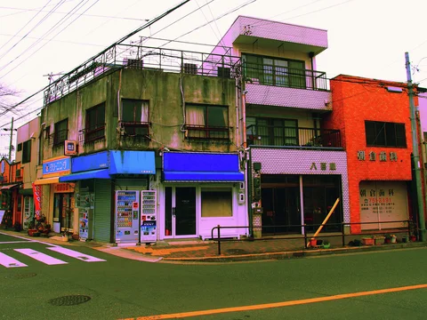 A weathered corner building with blue awnings stands beside narrow pink and orange facades beneath wires.