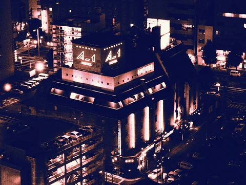 Bright vertical lights and rooftop hotel signs illuminate a dark building surrounded by streets and parked cars.