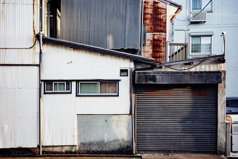 Small horizontal windows and a closed metal shutter punctuate a weathered corrugated building facade.
