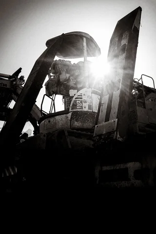 Sunlight flares through towering machinery parts silhouetted against a bright sky in black and white.