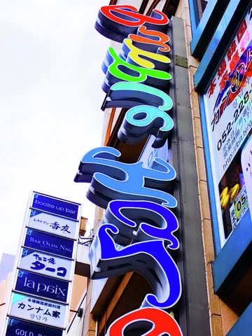 Large multicolored Japanese characters project vertically from a building beside smaller stacked business signs.