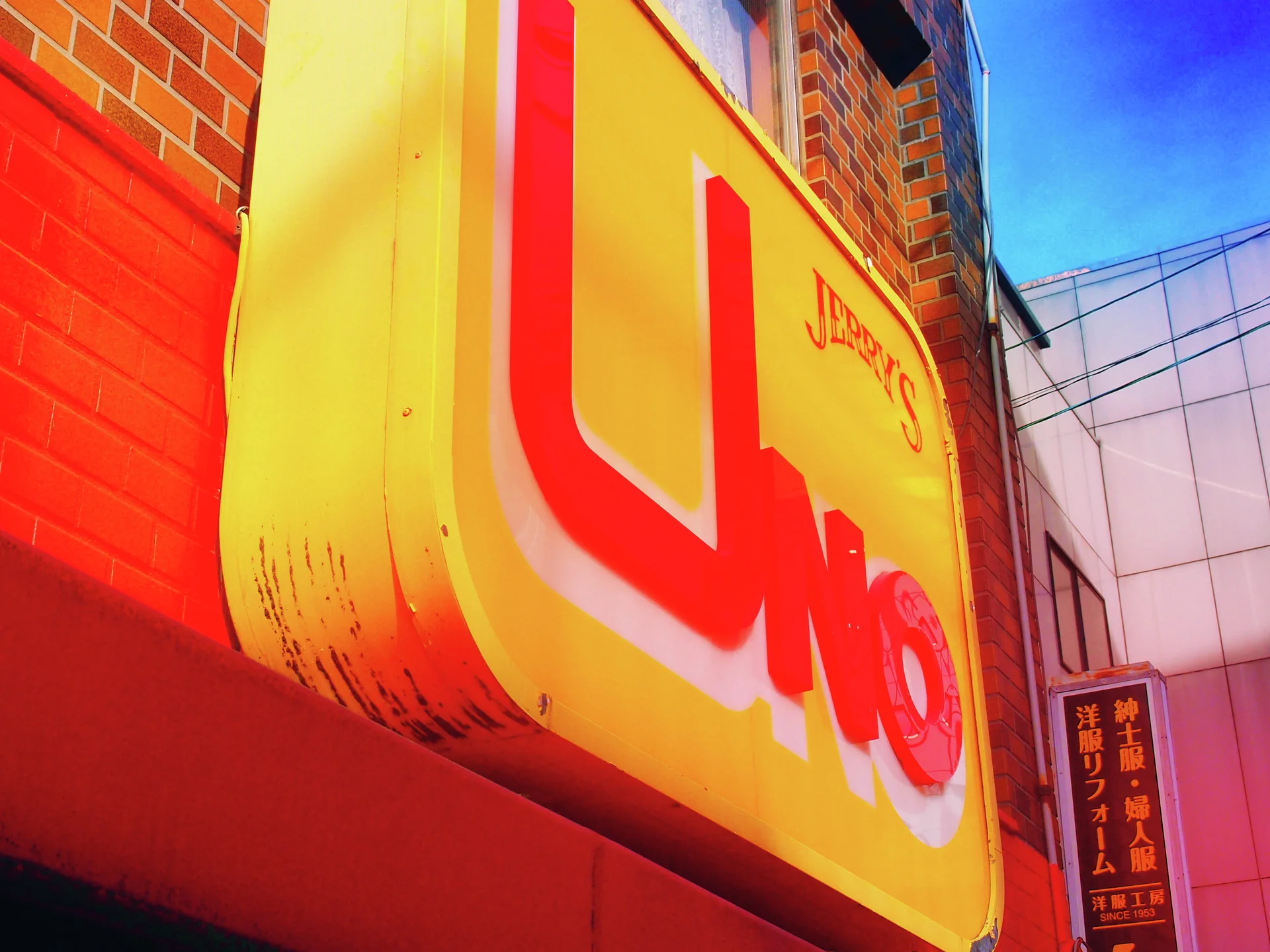 Large raised red Uno lettering covers a yellow projecting sign beside brickwork and a red wall.