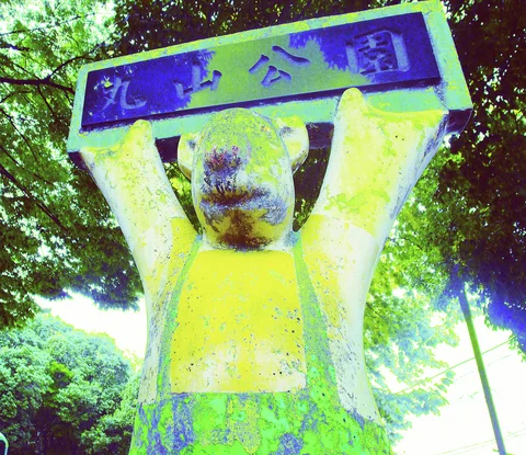A weathered bear statue raises a rectangular sign overhead beneath leafy trees.