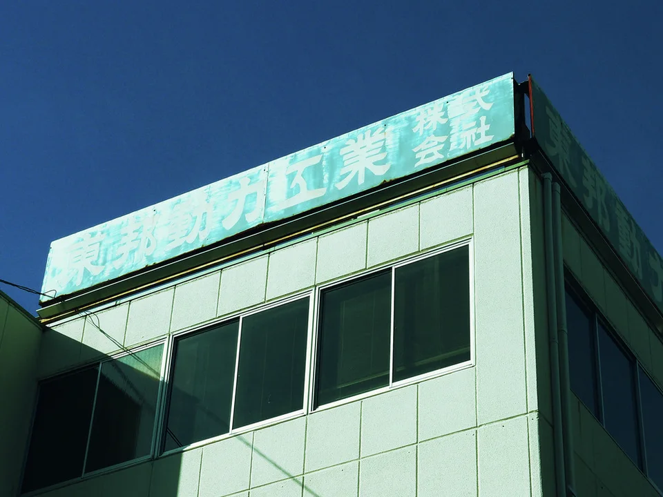 Faded white Japanese lettering spans a turquoise rooftop sign above dark windows and pale square facade panels.