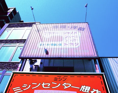 A faded pink sign with a sewing machine drawing rises above a red shop sign beneath blue sky.