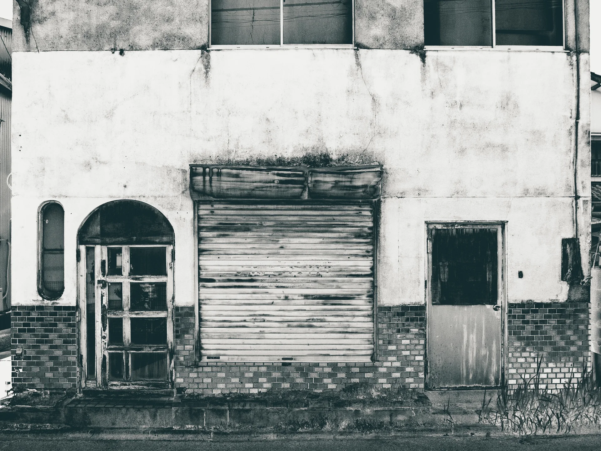 A stained facade has a closed metal shutter between mismatched narrow doors above dark brick trim.