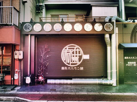 A closed brown restaurant shutter displays a white circular emblem beneath a row of round signs.