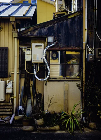 Utility boxes and looping pipes crowd a narrow wall beside a small window and potted greenery.