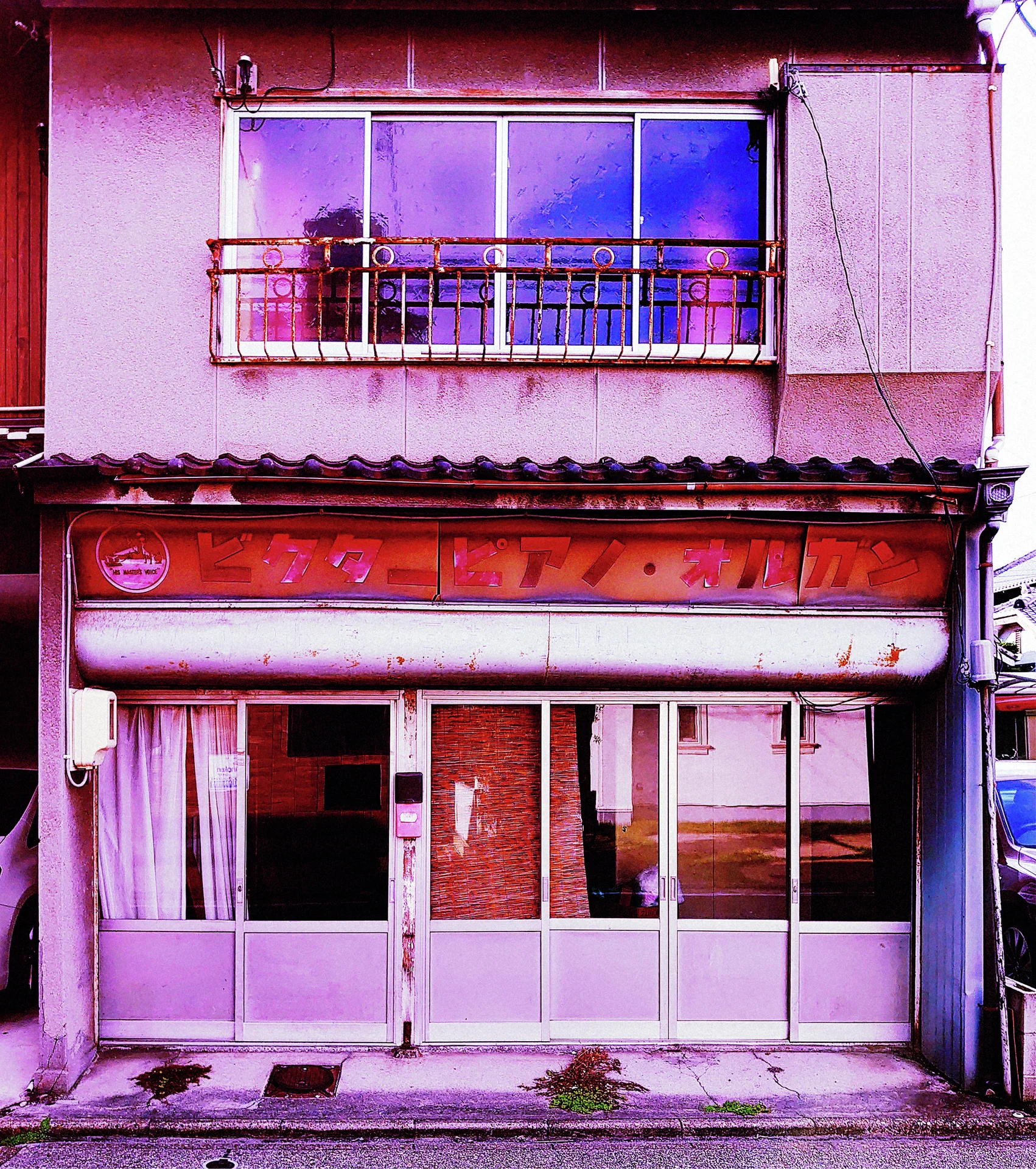 A faded pink storefront has closed sliding doors beneath a weathered sign and small upstairs balcony.