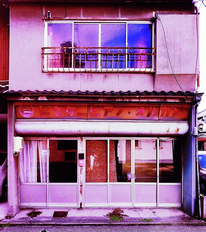 A faded pink storefront has closed sliding doors beneath a weathered sign and small upstairs balcony.