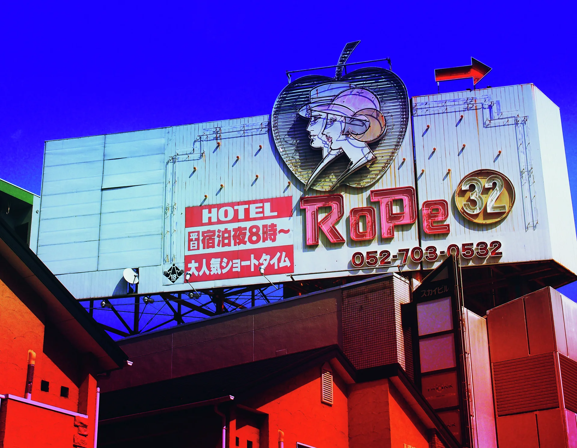 A large rooftop hotel sign combines red lettering, a profile illustration, and an arrow beneath blue sky.