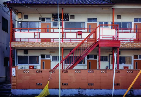 Bright red exterior stairs and railings connect two rows of doors on a small apartment building.