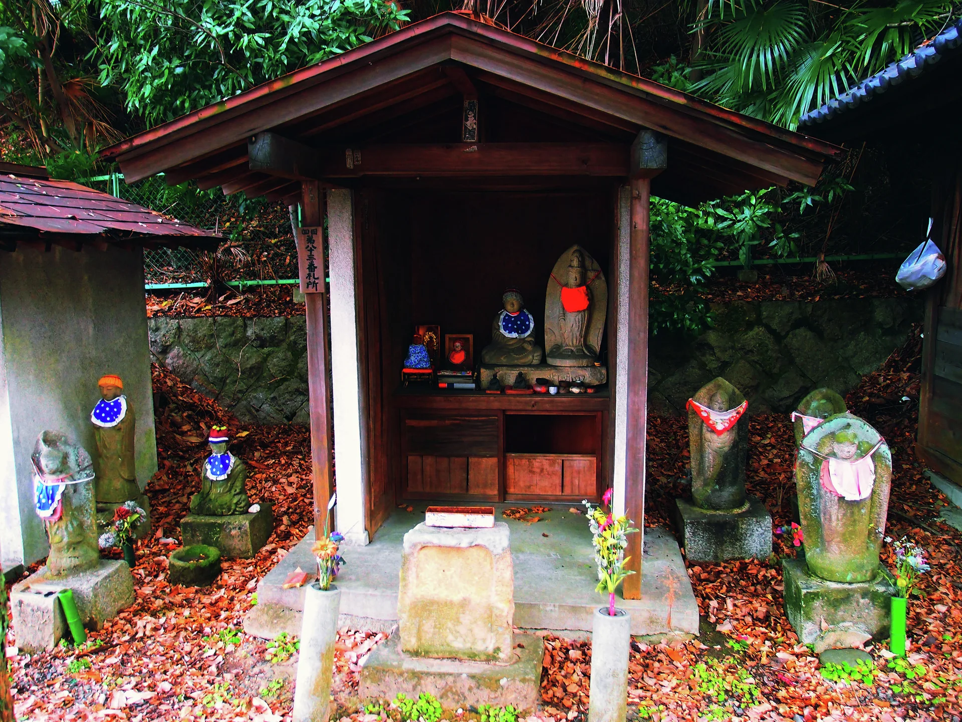 A small wooden shrine shelters stone figures surrounded by more statues and fallen leaves beneath green foliage.