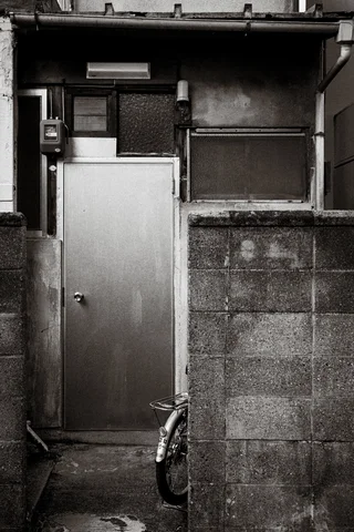 A narrow metal door stands behind a low block wall beside a bicycle in monochrome.