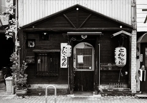 Two white paper lanterns flank the doorway of a dark wooden storefront in a black and white view.