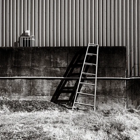 A ladder leans against a concrete wall below corrugated siding, casting a dark shadow in black and white.
