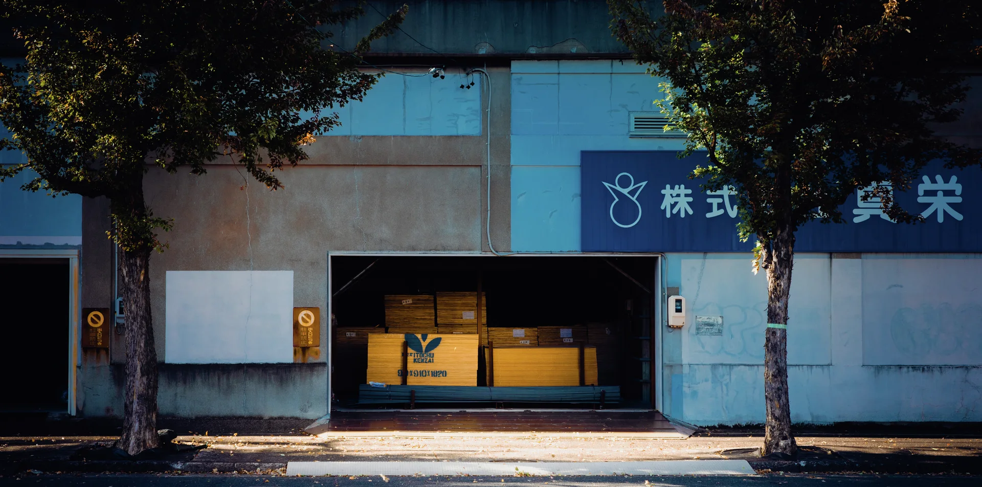 Trees frame a blue industrial facade with Japanese signage and an open bay containing yellow equipment.