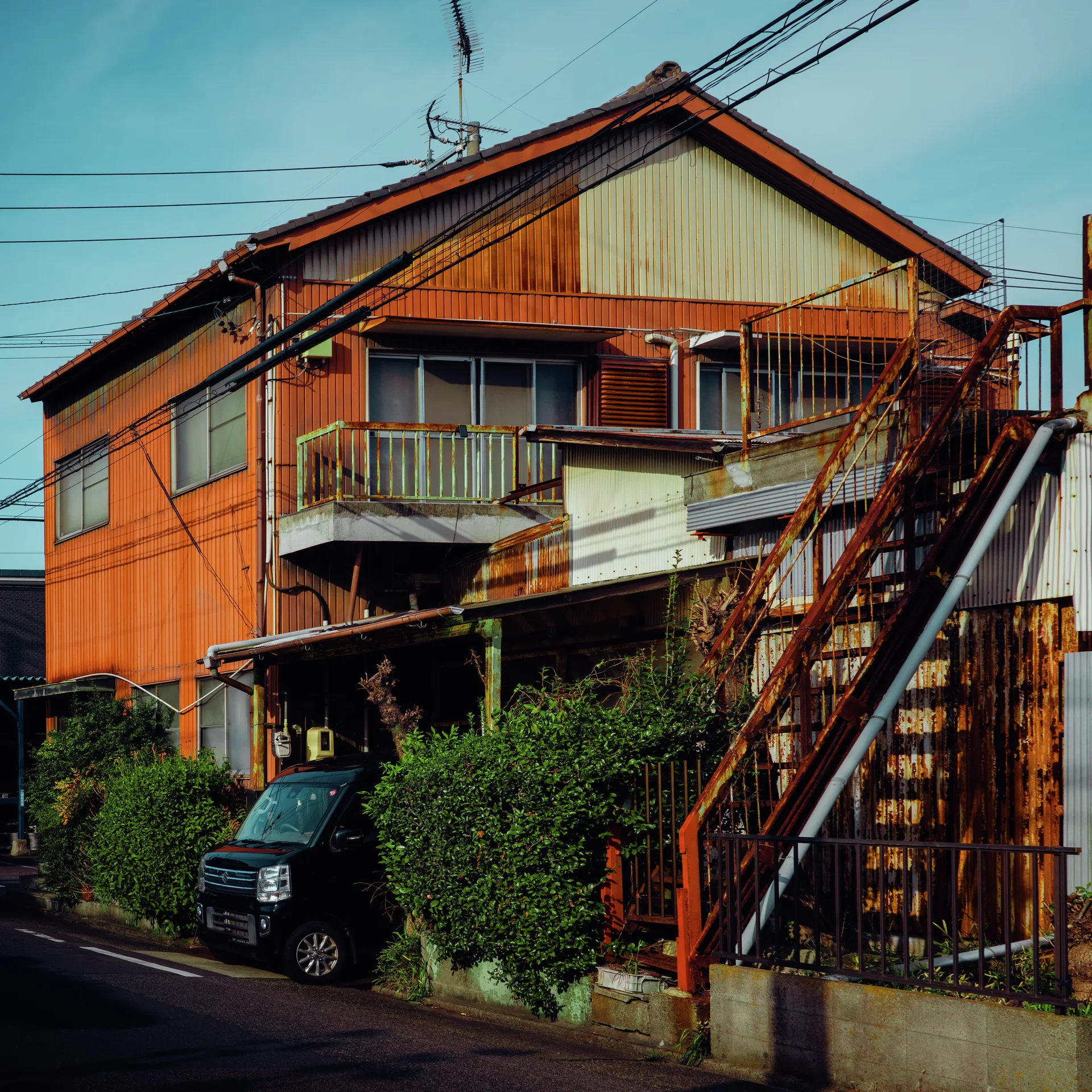 Orange siding and rusty outdoor stairs frame balconies above dense greenery and a parked dark van.