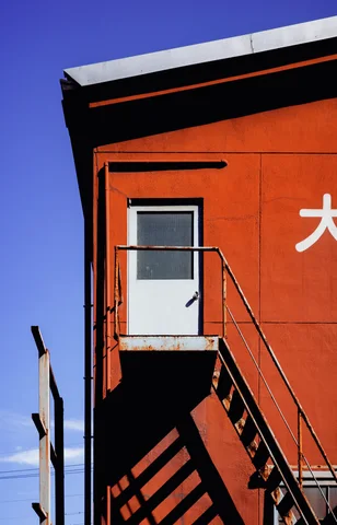 An exterior staircase casts sharp shadows across an orange wall beneath a small white door.