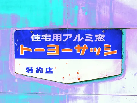 Orange and white Japanese lettering fills a blue-and-white shield-shaped sign on a pale reflective wall.
