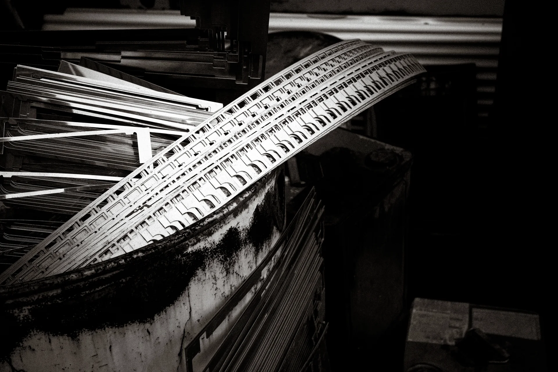 A patterned metal strip lies diagonally among stacked scrap pieces inside a dark workshop in black and white.