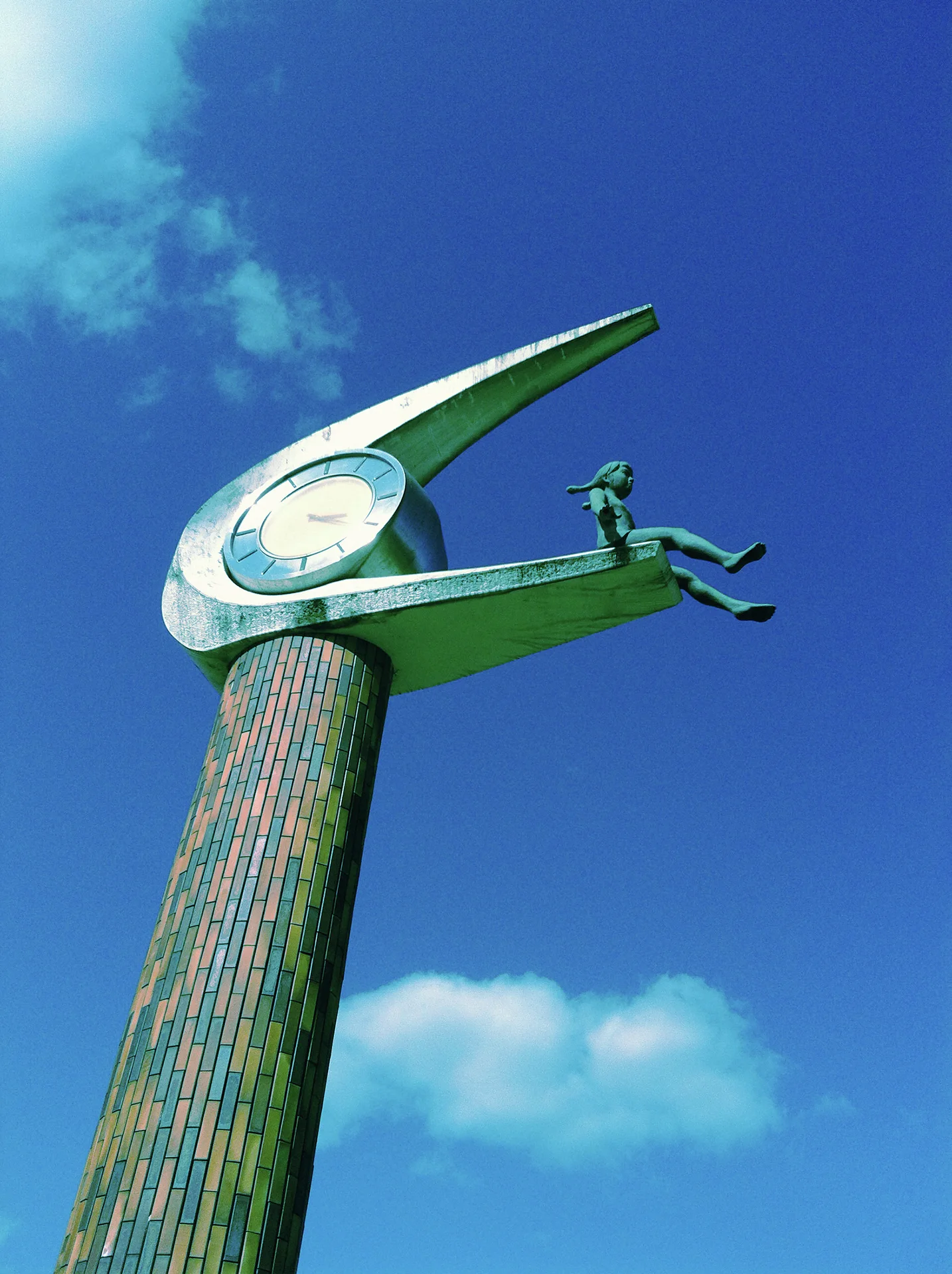 A wing-shaped clock with a small seated figure tops a textured column against a blue sky.