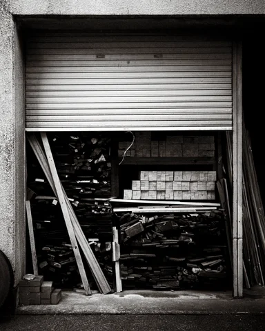A partly raised shutter reveals stacks of timber and blocks behind leaning boards in black and white.