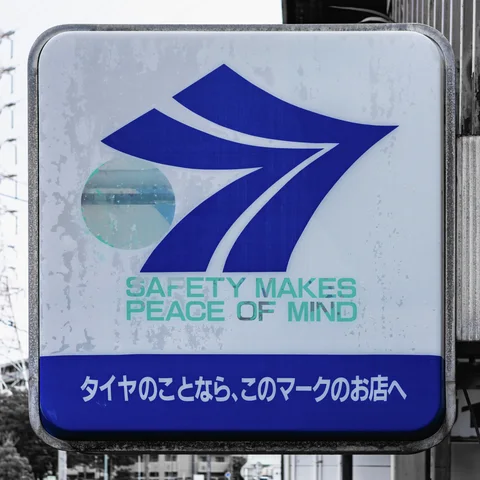 A weathered pale sign displays a large blue arrow-like emblem above green safety lettering.