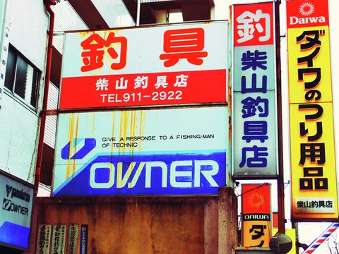 Red, blue, and yellow fishing tackle signs crowd a narrow building facade above a dark wooden entrance.