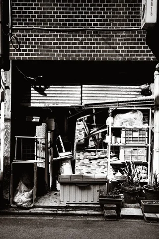 A cluttered open storefront contains stacked crates, frames, bags, and potted plants in black and white.