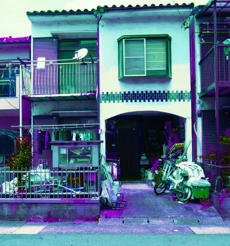 Bicycles and potted plants crowd an arched entrance beneath a narrow house balcony and curtained upper window.