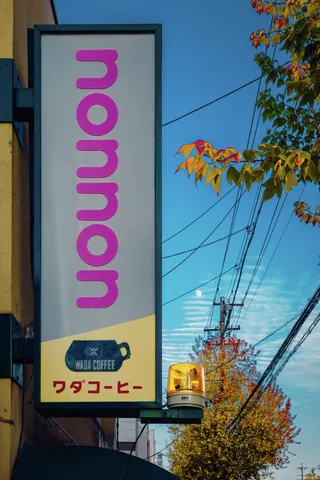 A slim pale sign with vertical pink lettering hangs beside autumn leaves against blue sky and utility wires.